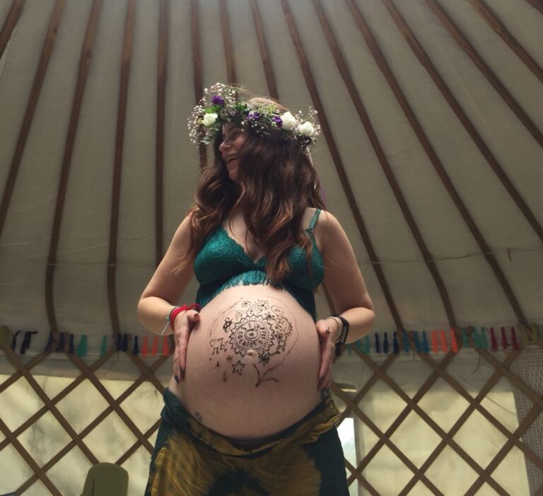 A pregnant doula standing with her pregnant bump painted with henna. A yurt is the background, in Horsham. Doula is wearing a flower crown.
